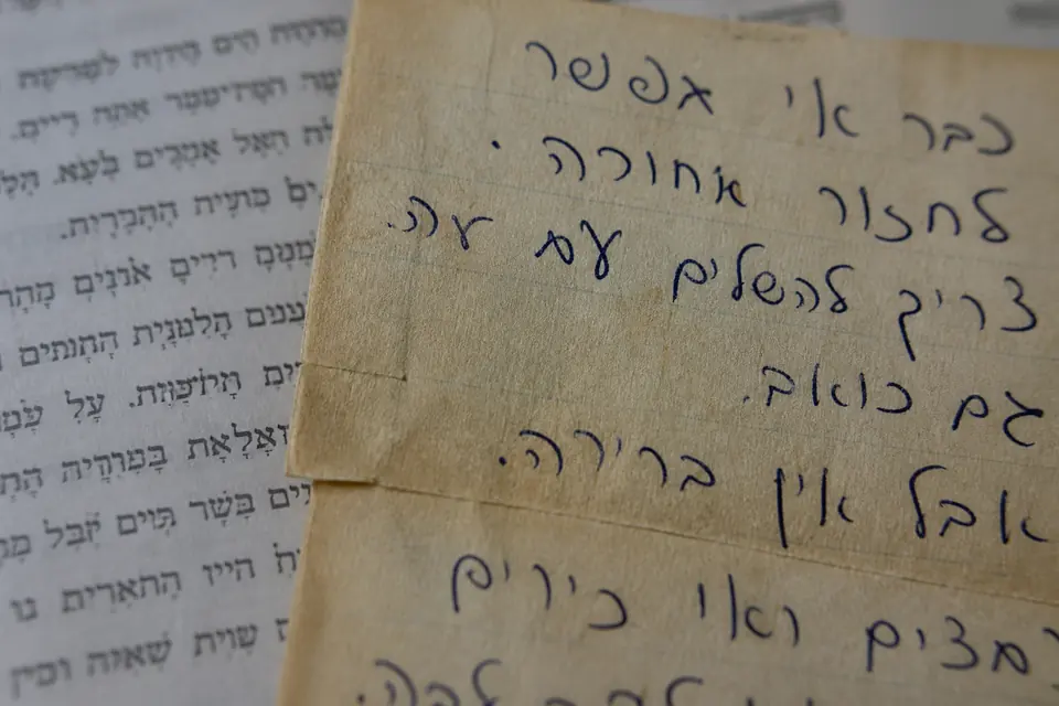 Close-up of messy Israeli cursive handwriting on paper, with a printed textbook letter out of focus behind it