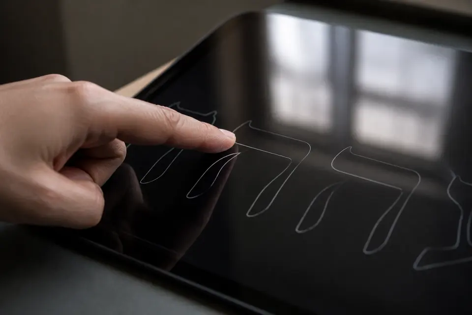 A finger tracing a pale Hebrew letter outline on tablet glass